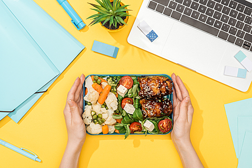 cropped view of woman holding lunch box with food near laptop with stationery