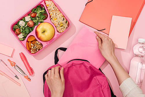 cropped view of woman packing backpack near lunch box with food and stationery