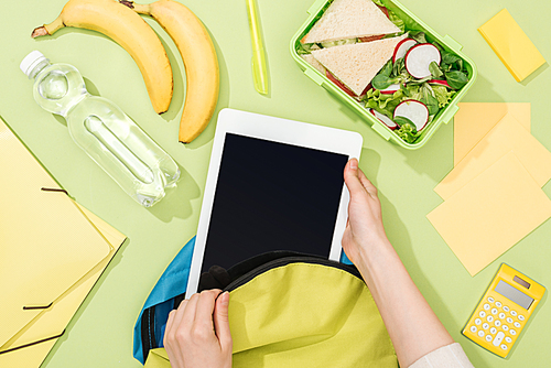 cropped view of woman packing digital tablet in backpack near lunch box, bananas, bottle of water and stationery