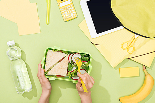 cropped view of woman hands with plastic utensils over lunch box with food near backpack, digital tablet, bottle of water and stationery