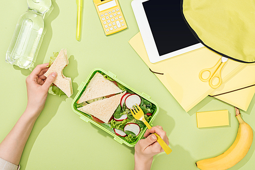 cropped view of woman hands with sandwich, plastic utensils over lunch box with food near backpack, digital tablet, bottle of water and stationery