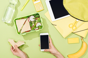 cropped view of woman hands with sandwich and smartphone near lunch box with food, backpack, digital tablet, bottle of water and stationery