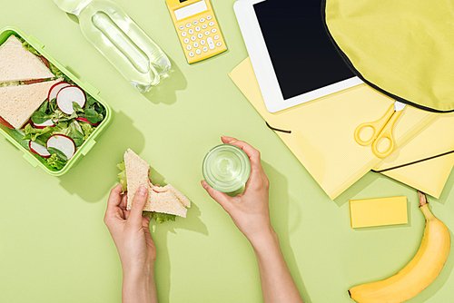 cropped view of woman hands with sandwich and glass of water near lunch box, backpack, digital tablet, bottle of water and stationery