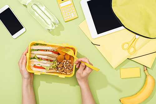 cropped view of woman hands with plastic utensils over lunch box with food near backpack, digital tablet, bottle of water and stationery