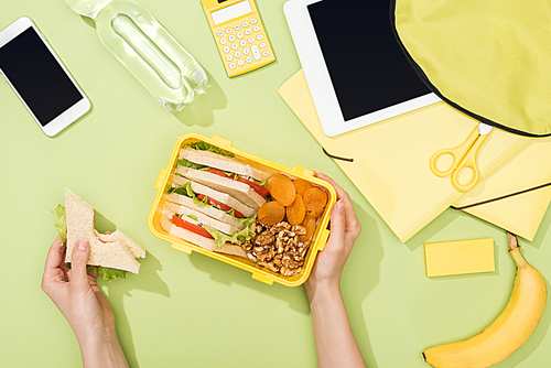 cropped view of woman hands with sandwich, plastic utensils over lunch box with food near backpack, digital tablet, bottle of water and stationery