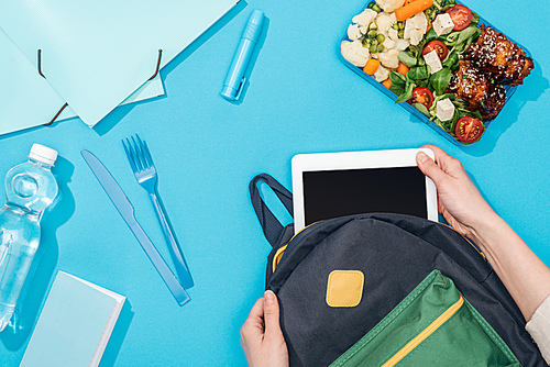 cropped view of woman packing digital tablet in backpack near lunch box, stationery and bottle of water