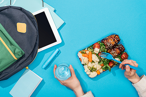 cropped view of woman holding lunch box and glass of water near backpack with folders, stationery and digital tablet