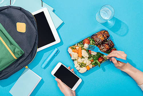 cropped view of woman hands with fork and smartphone near lunch box with food, backpack, digital tablet, glass of water and stationery