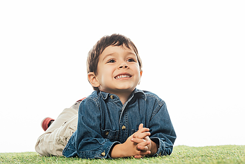 cute and smiling boy looking away and lying on grass isolated on white