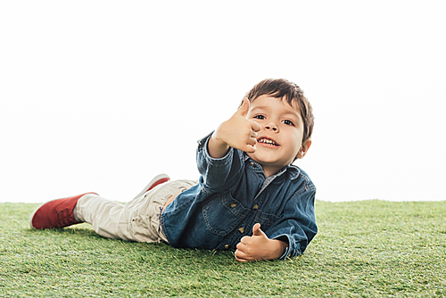 cute and smiling boy showing like and lying on grass isolated on white