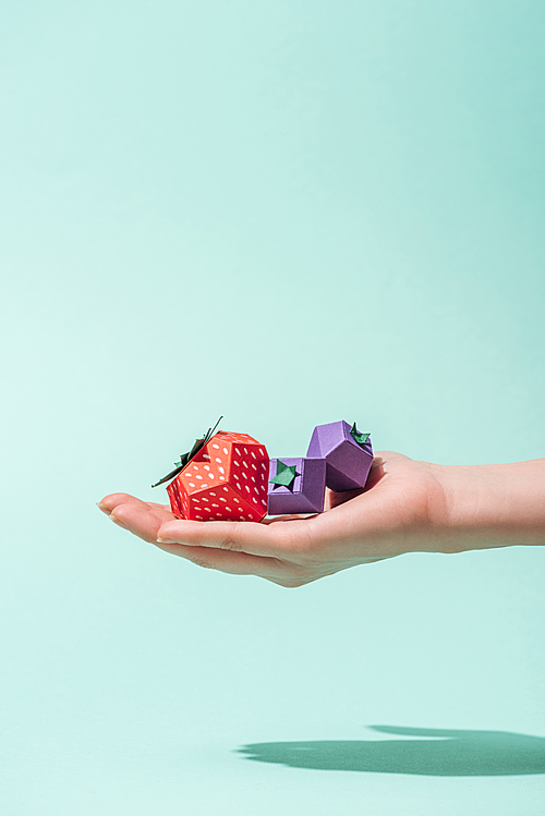 cropped view of young woman holding paper strawberry and blueberries on turquoise with copy space