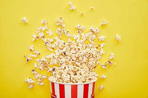 top view of delicious popcorn scattered from striped bucket on yellow background