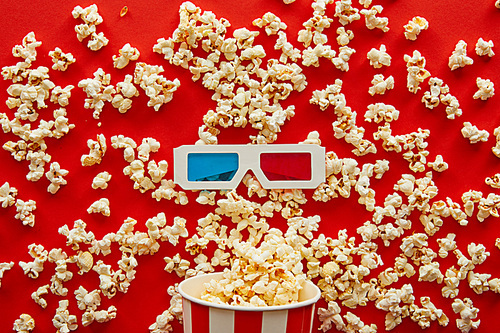 top view of delicious popcorn scattered from striped bucket near 3d glasses on red background