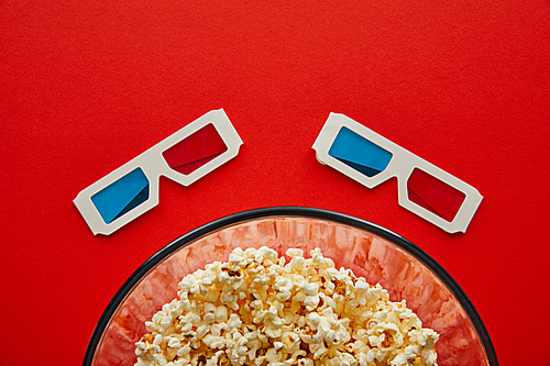 top view of delicious popcorn in glass bowl near 3d glasses isolated on red
