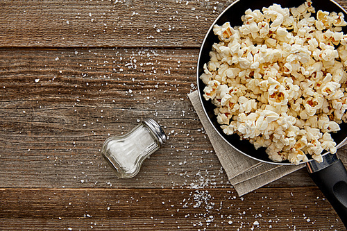 top view of delicious popcorn in frying pan near scattered salt on wooden background