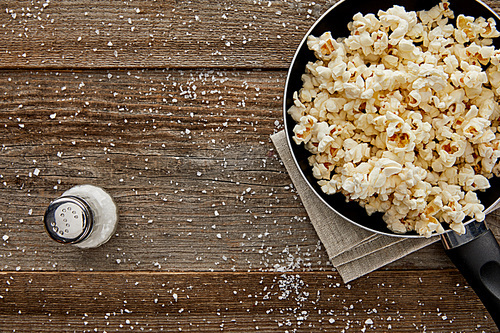 top view of fresh popcorn in frying pan with salt on wooden background