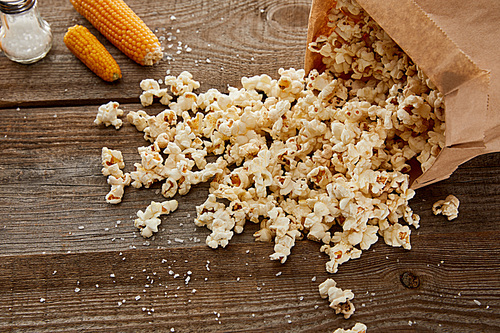 salty delicious popcorn scattered from paper bag near corn on wooden background