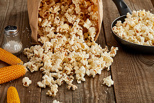 popcorn scattered from paper bag near corn and frying pan on wooden background