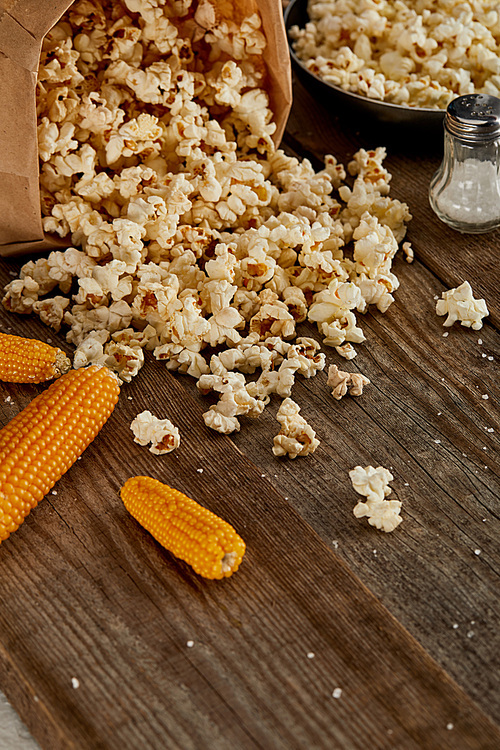 delicious popcorn scattered from paper bag near corn and frying pan on wooden background