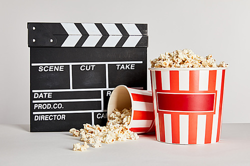 tasty popcorn in striped buckets near clapper board isolated on grey