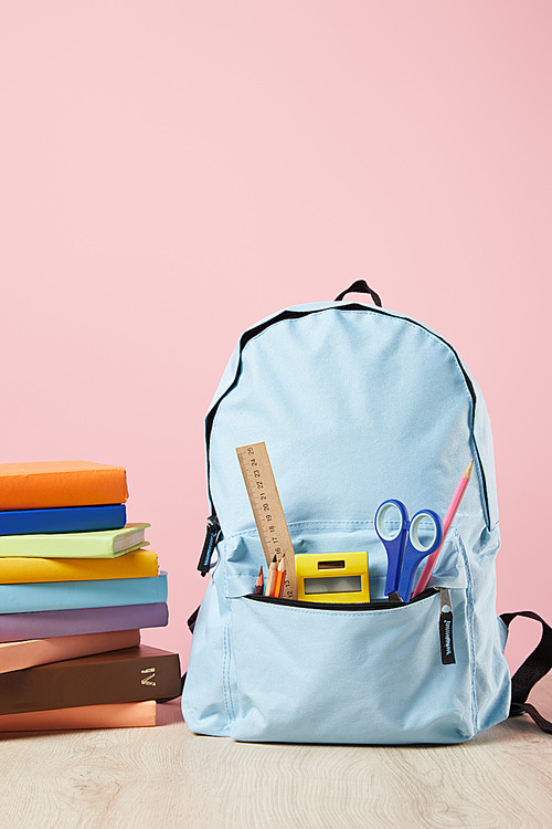 school blue backpack with supplies in pocket near stack of books isolated on pink