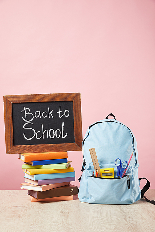 blue backpack with supplies in pocket near chalkboard with back to school lettering on books isolated on pink