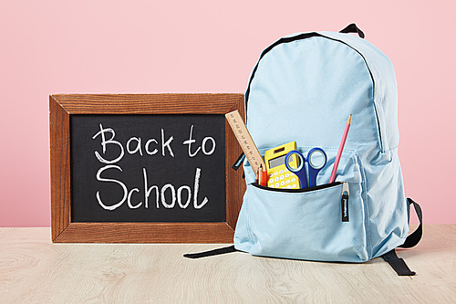 school blue backpack with supplies in pocket near chalkboard with back to school lettering isolated on pink