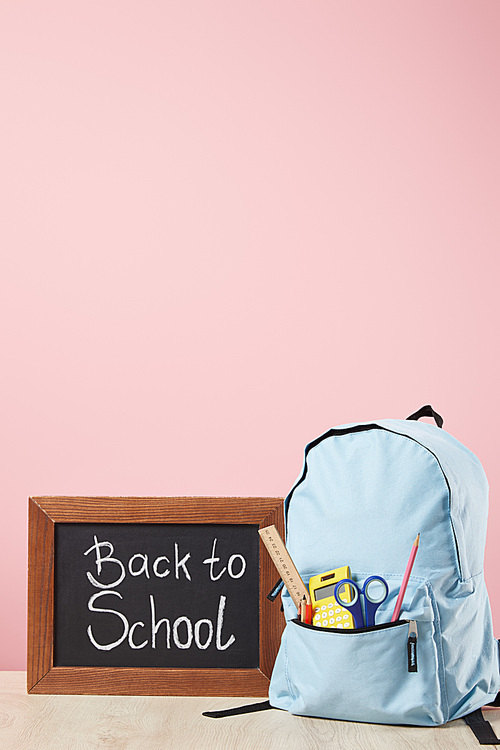 blue backpack with supplies in pocket near chalkboard with back to school lettering isolated on pink