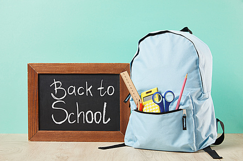 blue backpack with supplies in pocket near chalkboard with back to school lettering isolated on turquoise