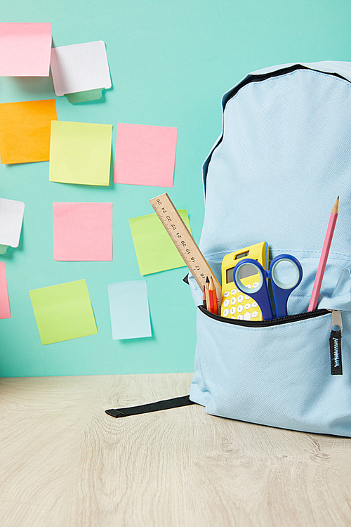 school blue backpack with supplies in pocket near multicolored sticky notes on turquoise wall