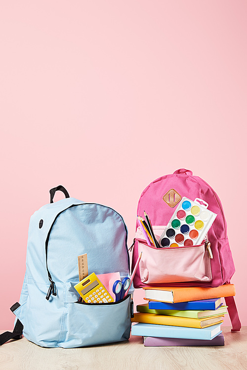 blue and pink backpacks with school supplies near stack of books isolated on pink
