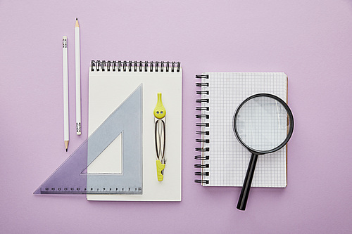 top view of ruler triangle and drawing compass on notebook near pencils and magnifier isolated on purple