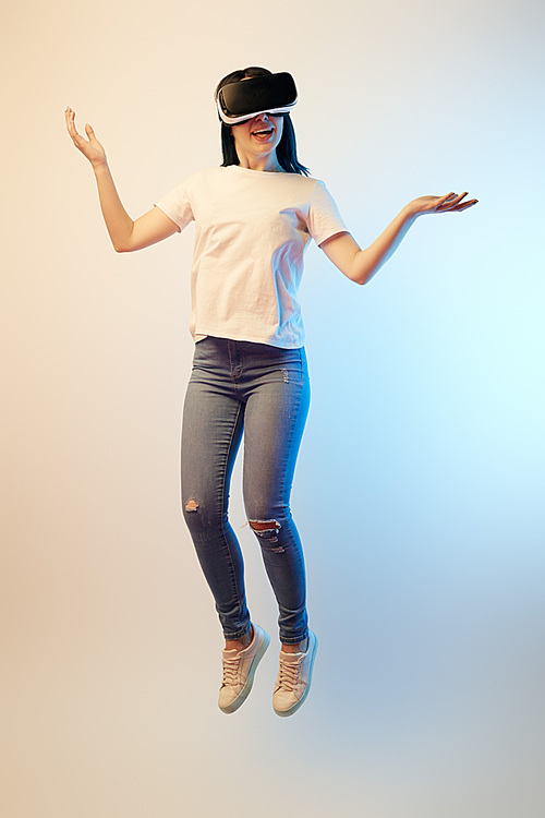 happy brunette woman in virtual reality headset levitating and showing shrug gesture on beige and blue