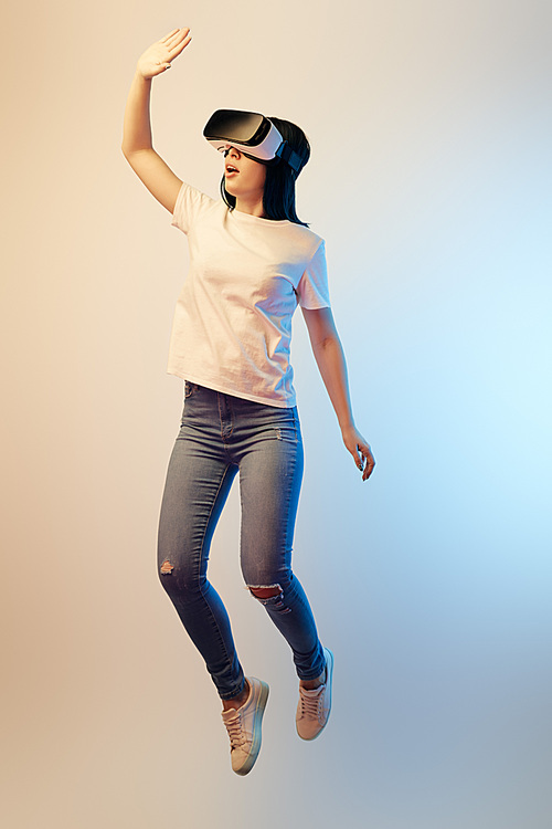 surprised brunette woman in virtual reality headset levitating and gesturing on beige and blue
