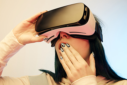 low angle view of surprised woman covering mouth while wearing virtual reality headset on beige and blue
