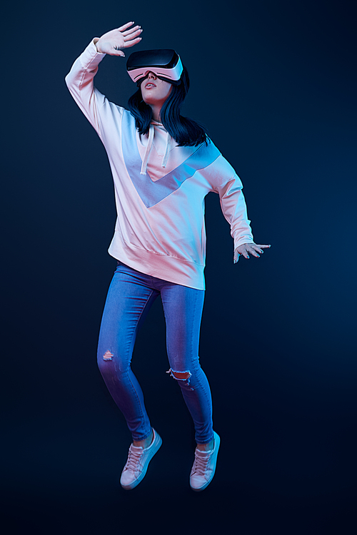 young woman gesturing while using virtual reality headset and jumping on blue