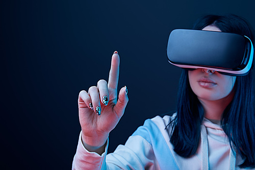 selective focus of young brunette woman wearing virtual reality headset and pointing with finger on blue