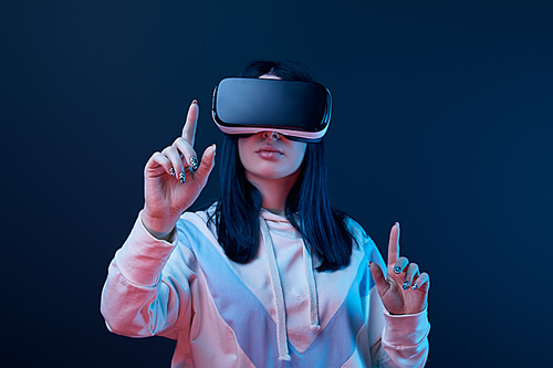 selective focus of young brunette woman wearing virtual reality headset and pointing with fingers on blue