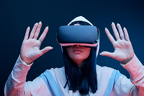 selective focus of brunette girl in hood gesturing while virtual reality headset on blue