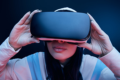 selective focus of brunette woman in hood touching virtual reality headset on blue