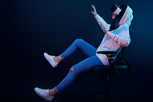 cheerful woman gesturing and using virtual reality headset while sitting on chair on blue