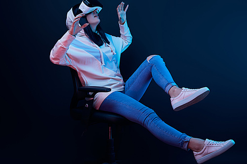surprised woman gesturing and using virtual reality headset while sitting on chair on blue