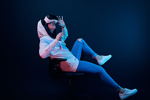 surprised girl gesturing and using virtual reality headset while sitting on chair on blue