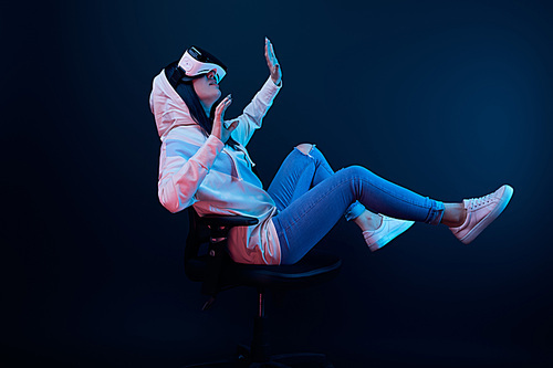 surprised young woman gesturing and using virtual reality headset while sitting on chair on blue
