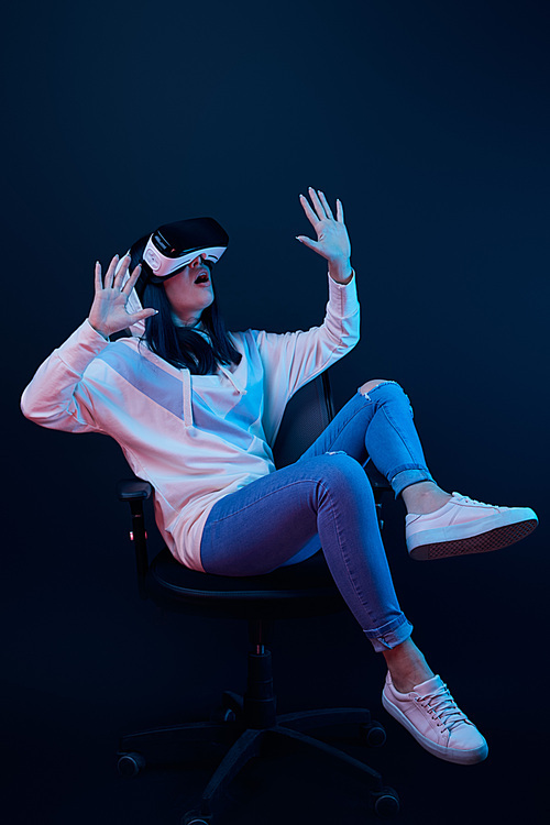 shocked young woman gesturing and using virtual reality headset while sitting on chair on blue