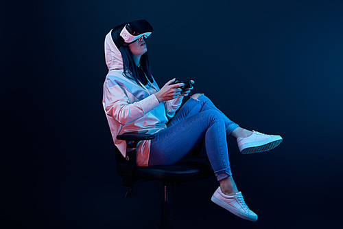 KYIV, UKRAINE - APRIL 5, 2019: Brunette woman sitting on chair and playing video game while wearing virtual reality headset on blue