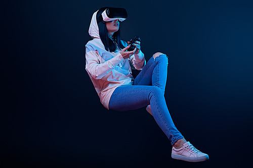 KYIV, UKRAINE - APRIL 5, 2019: Surprised young woman in virtual reality headset levitating and holding joystick on blue