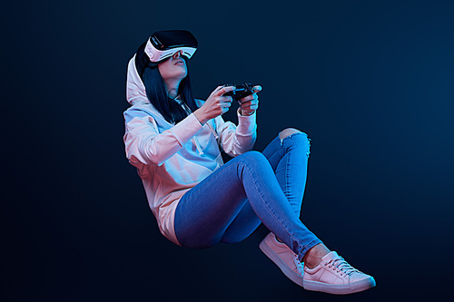 KYIV, UKRAINE - APRIL 5, 2019: Low angle view of young woman holding joystick while playing video game in virtual reality headset on blue
