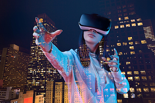 double exposure of brunette girl gesturing while wearing virtual reality headset and modern city with skyscrapers in nighttime
