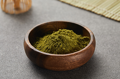 selective focus of green matcha powder in wooden bowl near bamboo whisk and table mat on grey table